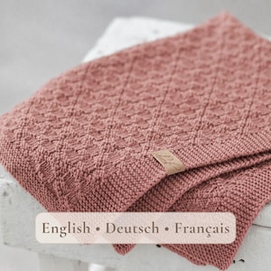 Può includere: Una coperta lavorata a maglia color rosa polvere con un motivo a rombi testurizzato sulla superficie. La coperta presenta un bordo in maglia unita e una piccola etichetta rettangolare color beige con il testo in corsivo English • Deutsch • Français.