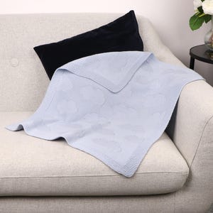 May include: A light blue knitted blanket featuring a repeating cloud pattern and a textured border, paired with a solid black rectangular velvet pillow. The blanket has a soft, woven appearance with a consistent knit structure throughout the fabric.