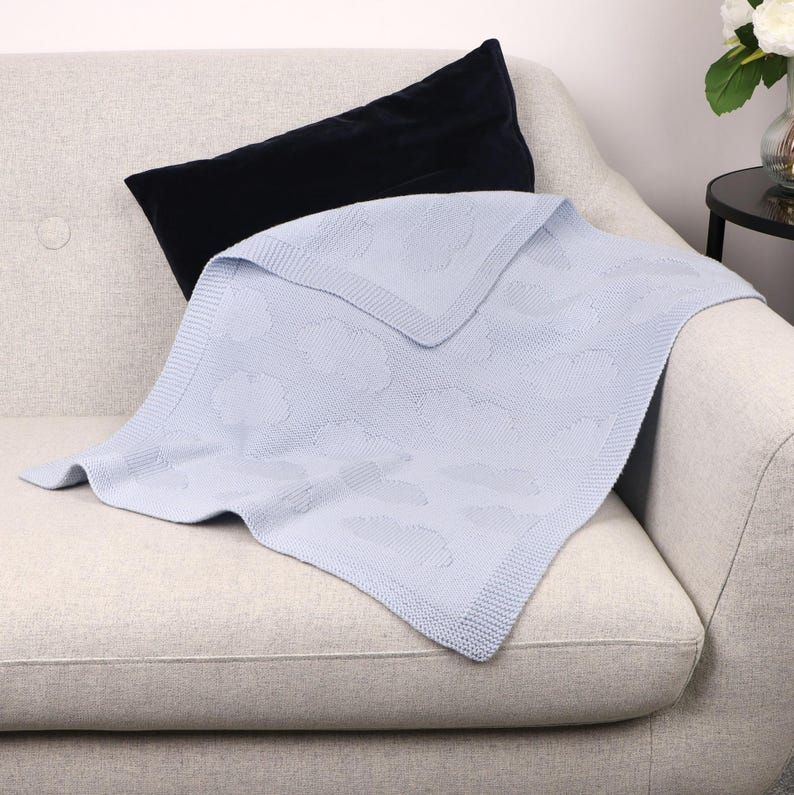 May include: A light blue knitted blanket featuring a repeating cloud pattern and a textured border, paired with a solid black rectangular velvet pillow. The blanket has a soft, woven appearance with a consistent knit structure throughout the fabric.