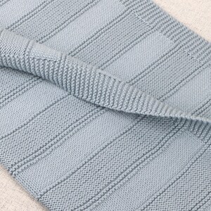 May include: A light blue knitted blanket featuring a textured pattern of vertical garter stitch stripes alternating with smooth stockinette stitch panels. The fabric has a folded edge, revealing the consistent knit construction and soft blue yarn.