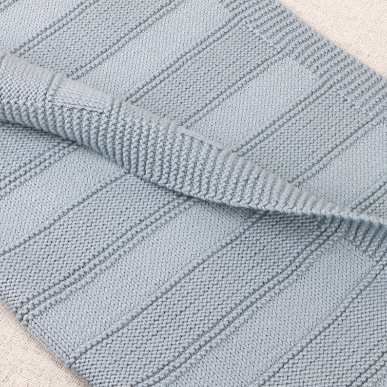 May include: A light blue knitted blanket featuring a textured pattern of vertical garter stitch stripes alternating with smooth stockinette stitch panels. The fabric has a folded edge, revealing the consistent knit construction and soft blue yarn.