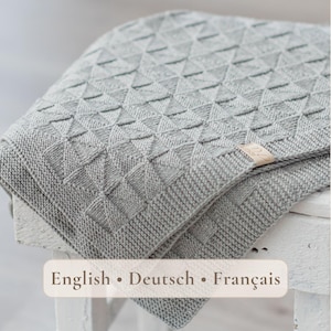 Könnte beinhalten: Eine hellgraue Strickdecke mit einem sich wiederholenden geometrischen Dreiecksmuster und einem strukturierten Rand. Ein kleines rechteckiges beiges Stoffetikett ist am Rand der Decke befestigt. Die Decke liegt gefaltet auf einer weißen Holzoberfläche.