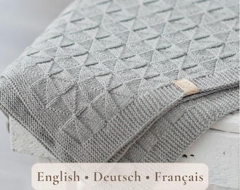 May include: A light gray knitted blanket featuring a repeating geometric triangle pattern and a textured border. A small rectangular beige fabric tag is attached to the edge of the blanket. The blanket is folded and draped over a white wooden surface.