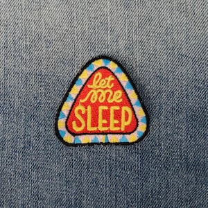 Könnte beinhalten: Ein roter, gelber und blauer dreieckiger Patch mit dem Text "let me sleep", der darauf gestickt ist. Der Patch ist auf eine blaue Jeansjacke genäht.