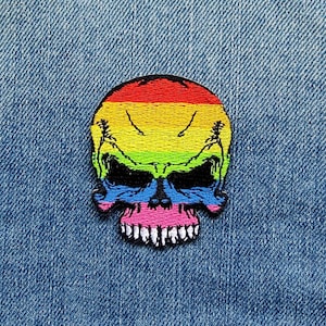 Könnte beinhalten: Ein Regenbogenfarbener Totenkopf-Patch mit schwarzem Umriss. Der Totenkopf ist auf einem Jeansstoff gestickt.