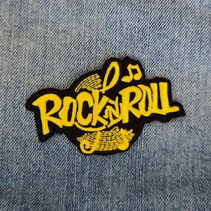 Può includere: Testo giallo "Rock 'n' Roll" con contorno nero su una grafica di disco in vinile nero. La grafica è circondata da un contorno nero e ha note musicali in alto.