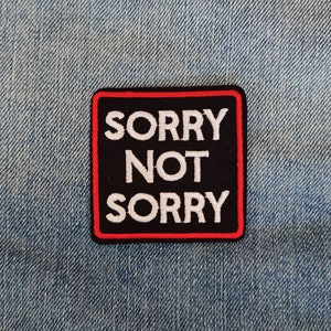 Puede incluir: Un parche bordado en blanco y negro con costuras rojas. El parche es rectangular y dice "SORRY NOT SORRY" en letras blancas sobre un fondo negro.