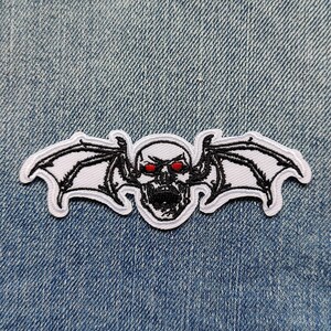 May include: White embroidered patch of a skull with red eyes and bat wings on a blue denim background.