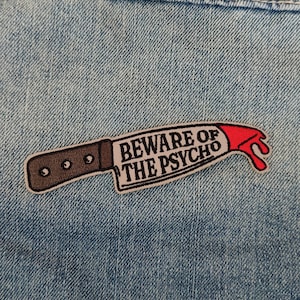 Peut inclure: Un patch brodé rouge et noir avec un couteau et le texte "Beware of the Psycho".