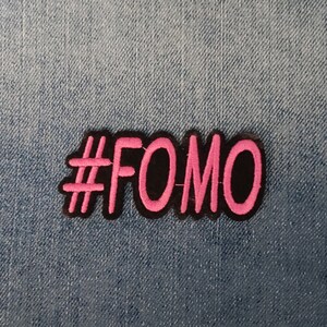 Op de afbeelding: Een roze en zwarte geborduurde patch met de tekst "#FOMO" op een denim achtergrond.