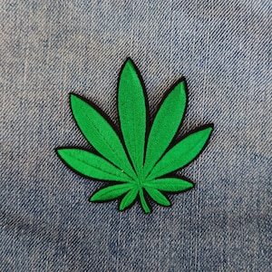 Può includere: Toppa a forma di foglia di cannabis verde con bordi neri, su sfondo denim. La toppa ricamata mostra le venature e i bordi della foglia in dettaglio. Ideale per personalizzare giacche, borse o altri articoli in tessuto.