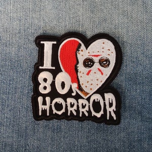 Op de afbeelding: Een geborduurde patch in zwart en wit met een rood hart. Het hart heeft een wit hockeymasker met een rode streep en zwarte ogen. De tekst "I 80's HORROR" is in wit op een zwarte achtergrond geschreven.