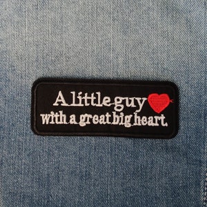 Könnte beinhalten: Schwarzer gestickter Aufnäher mit weißem Text, der "A little guy with a great big heart" lautet, und einem roten Herzen.