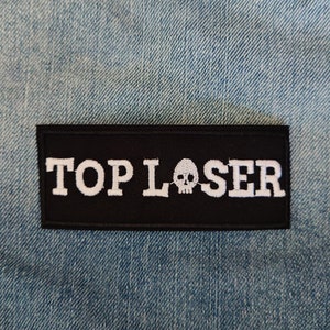 May include: Black embroidered patch with white text that reads "TOP LOSER" and a white skull graphic.