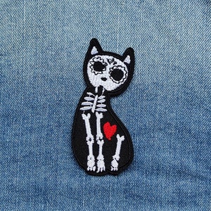 Peut inclure: Patch thermocollant brodé noir représentant un chat squelette avec un cœur rouge sur un fond en jean.
