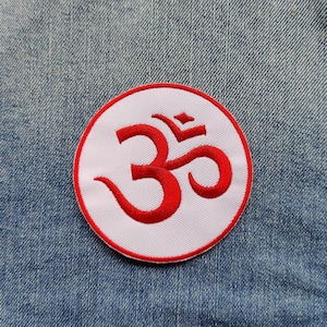 May include: A white circular patch with a red embroidered Om symbol. The patch is sewn onto a blue denim jacket.