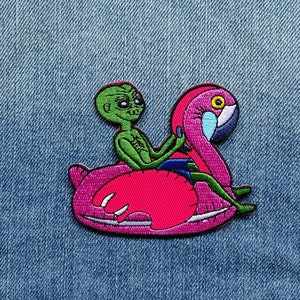 Peut inclure: Écusson brodé représentant un extraterrestre vert sur une bouée gonflable flamant rose. L'extraterrestre porte un short bleu. Le flamant rose a un œil jaune et des détails bleus et blancs. Sur fond de denim bleu.