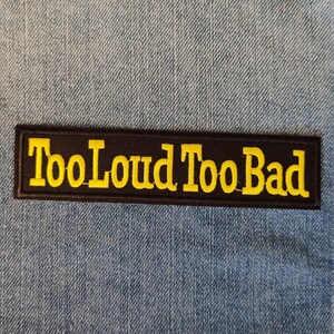 Könnte beinhalten: Ein gestickter schwarzer und gelber Aufnäher mit dem Text "Too Loud Too Bad" auf einem Jeansstoff.