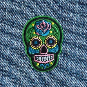 May include: Green embroidered sugar skull patch with a pink and blue flower, yellow and blue eyes, and a pink and white smile.