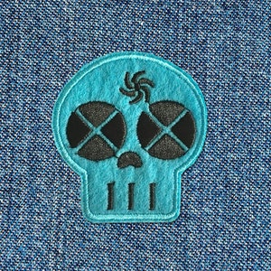 Può includere: Una toppa a forma di teschio blu turchese con occhi ricamati neri e un vortice nero sulla fronte. La toppa è su uno sfondo di denim blu.