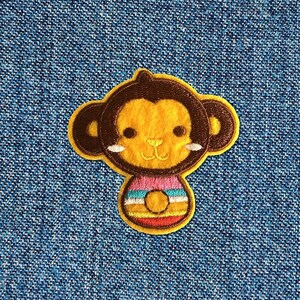 May include: A brown and yellow embroidered monkey patch with a pink and yellow striped shirt. The monkey has a brown nose and brown eyes.
