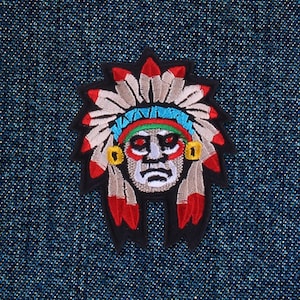 Peut inclure: Écusson brodé représentant le visage d'un chef amérindien avec une coiffe de plumes. L'écusson a une bordure noire et est posé sur un fond en denim bleu. La coiffe comprend des accents rouges, blancs et bleus.