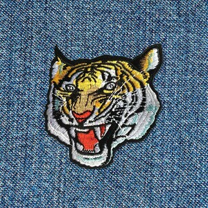 Può includere: Un toppa ricamata con la testa di una tigre. La tigre ha strisce gialle e nere, un muso bianco e una bocca rossa aperta.