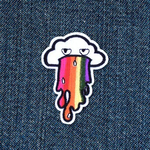 May include: A white cloud with a frowning face is vomiting a rainbow on a blue denim background.