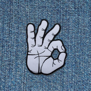 Könnte beinhalten: Weißer gestickter Aufnäher einer Hand, die das OK-Zeichen macht. Die Hand ist schwarz umrandet.