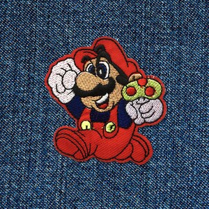 Könnte beinhalten: Gestickter Aufnäher mit einer Comicfigur, Mario, in einem roten Overall und einer roten Mütze, der einen grünen Pilz hält. Der Aufnäher befindet sich auf einem blauen Jeansstoff.