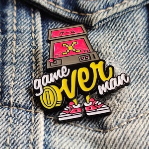 May include: A black and yellow enamel pin with a retro arcade game, the words "game over man" and a pair of pink sneakers. The pin is attached to a blue denim jacket.