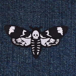 Könnte beinhalten: Schwarz-weiß gestickter Patch einer Motte mit einem Totenkopf auf ihrem Körper. Die Motte befindet sich auf einem blauen Jeansstoff.