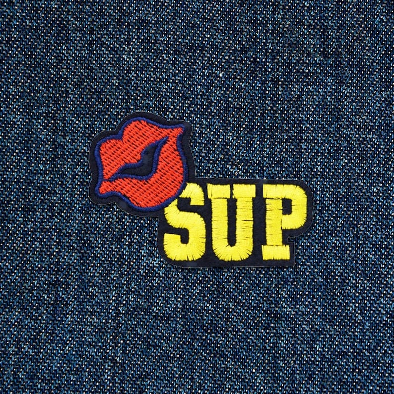 May include: Embroidered patch with a red lip design and blue outline, alongside the word "SUP" in yellow block letters. The patch is set against a dark blue denim background.