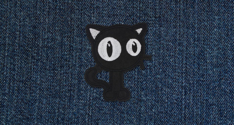 Op de afbeelding: Zwarte geborduurde kattenpatch met witte ogen. De patch is op een blauwe denim achtergrond.