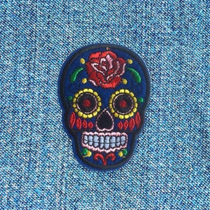 May include: Embroidered patch with a sugar skull design. The skull is blue with red, yellow, and green detailing. A red rose is embroidered on the top. The patch is set against a blue fabric background.