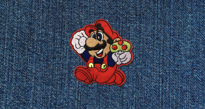 Op de afbeelding: Gestikte patch met een cartoonfiguur, Mario, in een rood pak en een rode muts, die een groene paddenstoel vasthoudt. De patch is op een blauwe denim achtergrond.