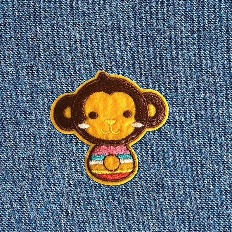 Pu&ograve; includere: Una toppa di scimmia ricamata in marrone e giallo con una camicia a righe rosa e gialla. La scimmia ha un naso marrone e occhi marroni.