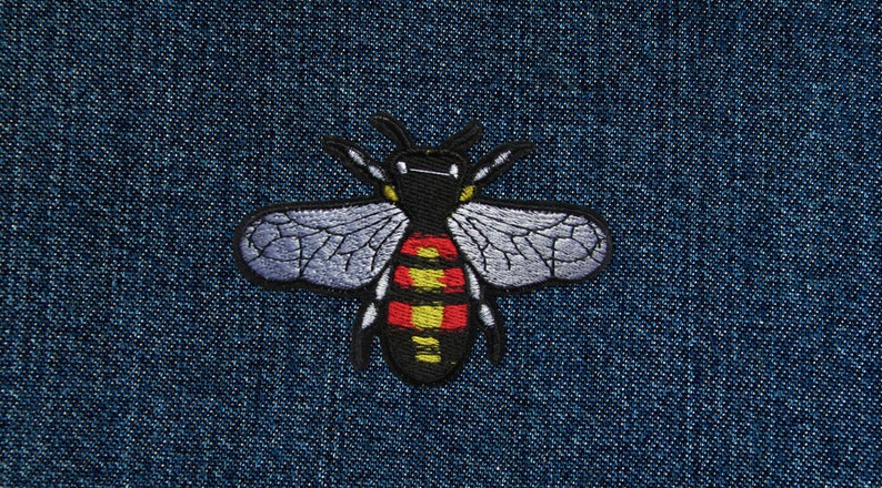 Peut inclure: Patch brod&eacute; en forme d'abeille avec des ailes noires, un abdomen ray&eacute; jaune et rouge et une t&ecirc;te noire. Le patch est sur un fond en jean bleu.