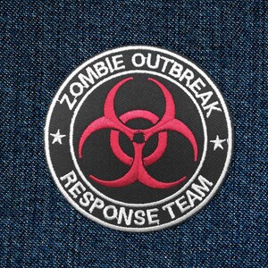 Peut inclure: Un écusson circulaire noir et blanc avec le texte "Zombie Outbreak Response Team" en lettres blanches autour d'un symbole de biohazard rouge.