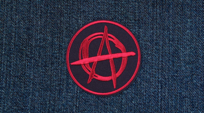 Puede incluir: Un parche circular negro con un s&iacute;mbolo de anarqu&iacute;a rojo bordado. El parche est&aacute; cosido sobre una tela vaquera azul.