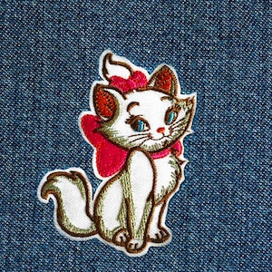 May include: White embroidered cat with a pink bow, sitting on a blue denim background.