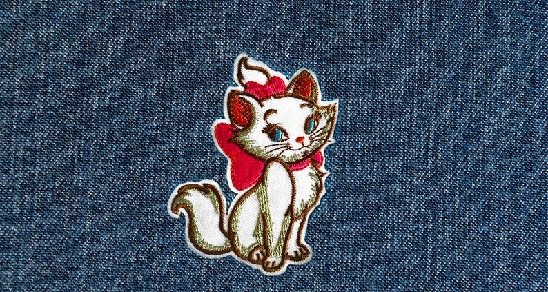 K&ouml;nnte beinhalten: Wei&szlig; gestickter Kater mit einer rosa Schleife, der auf einem blauen Jeansstoff sitzt.