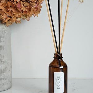 May include: A brown glass bottle diffuser with black and natural reed sticks. The bottle has a white label with the text "BOUTIQUE DOUZE HOME".