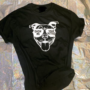 以下が含まれることがあります： 黒いTシャツに、サングラスをかけたピットブルの白いグラフィックがプリントされています。サングラスには「Show Me Your Pitties」という文字が書かれています。