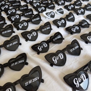 May include: A collection of heart-shaped sunglasses in black, with the initials "G&D" and the date "27.07.2024" printed in white. Some sunglasses are white. The sunglasses are arranged on a white surface.