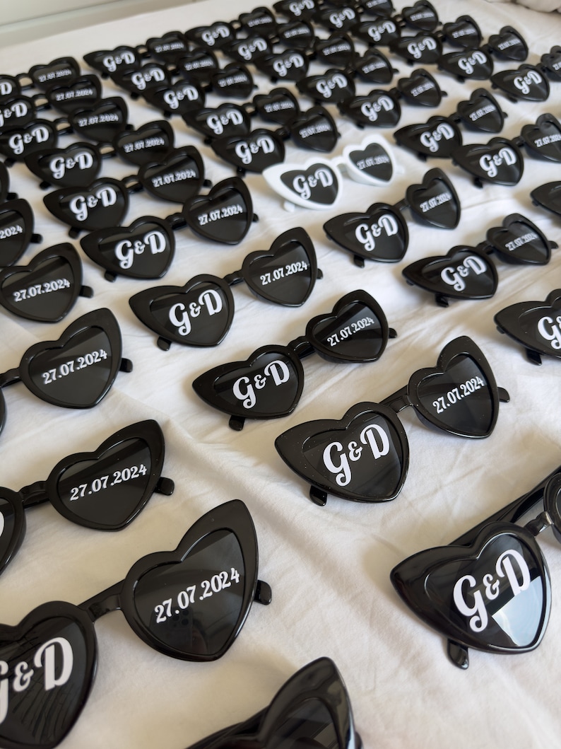 May include: A collection of heart-shaped sunglasses in black, with the initials "G&D" and the date "27.07.2024" printed in white. Some sunglasses are white. The sunglasses are arranged on a white surface.