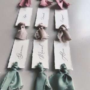 May include: Nine place cards with handwritten names in elegant calligraphy. Each card is adorned with a silk ribbon in shades of pink, beige, and green. The cards are arranged on a gray surface, ideal for weddings or special events.
