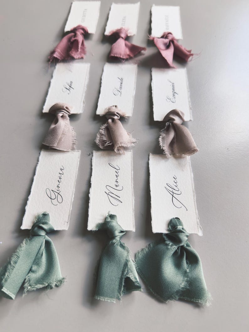 May include: Nine place cards with handwritten names in elegant calligraphy. Each card is adorned with a silk ribbon in shades of pink, beige, and green. The cards are arranged on a gray surface, ideal for weddings or special events.