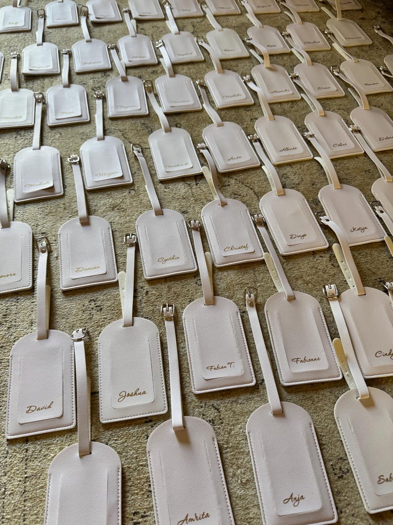 May include: Numerous light pink luggage tags with silver-coloured hardware and matching straps. Each tag features a rectangular window and is personalised with a name in gold lettering. The tags are arranged on a textured surface.