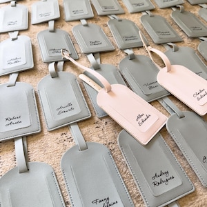 May include: A selection of personalised luggage tags in shades of grey and one in pink. Each tag has a name written in script. The tags are made of a smooth material and have a strap for attachment.